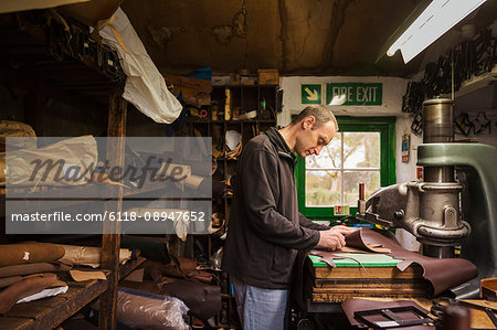Man standing in a shoemaker's workshop, cutting brown leather.