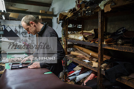 Man standing in a shoemaker's workshop, cutting brown leather.