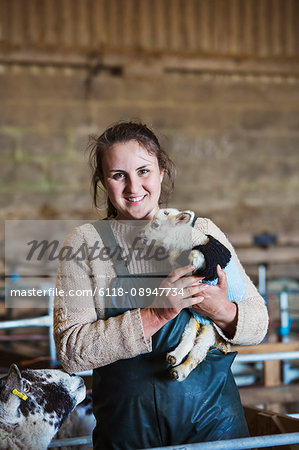Woman standing in a barn, holding a newborn lamb dressed in a knitted jumper.