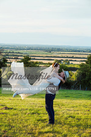 Newlyweds standing in a grass meadow overlooking a rural view, groom hugging and swinging the bride in the air.