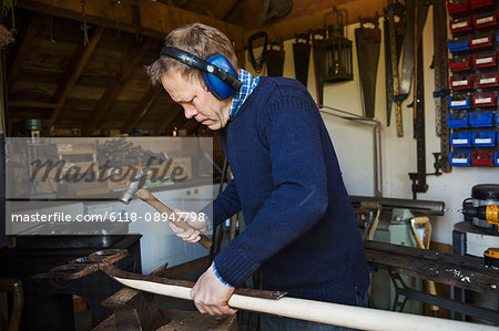 Man standing in a workshop, wearing ear protectors, holding a hammer, working on a piece of wood.