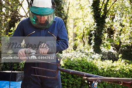 Man standing outdoors, wearing a face mask, working on a large metal garden fork or pitchfork with an angle grinder.