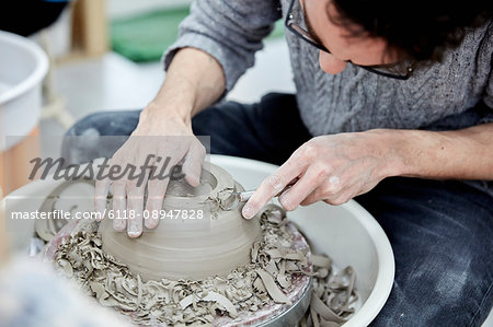 A man seated at a potter's wheel working and shaping a clay pot by removing excess clay.