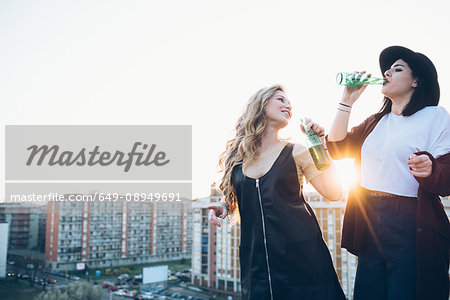 Two young women, on roof top, drinking bottled beer