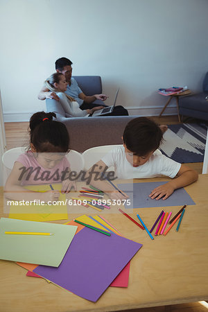 Boy and girl drawing in paper while parents using laptop in background