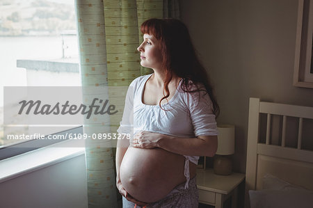 Thoughtful pregnant woman looking through window in bedroom
