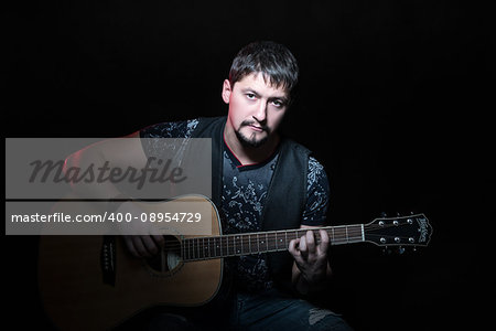 Portrait of handsome bearded man sitting in a chair with an acoustic guitar in hand, isolated on a black background