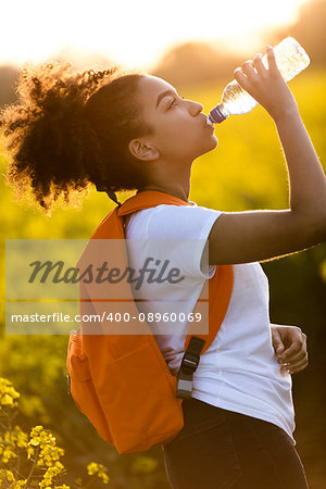 Outdoor portrait of beautiful happy mixed race African American girl teenager female young woman drinking water from a bottle in a field of yellow flowers at sunset in golden evening sunshine