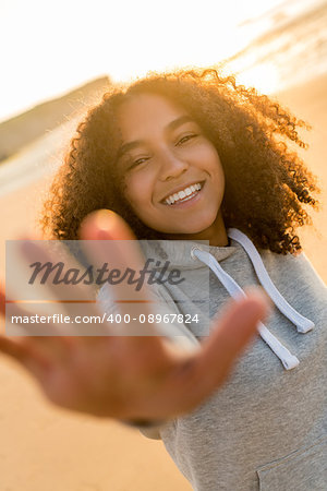 Outdoor portrait of beautiful happy mixed race African American girl teenager female young woman on a beach reaching out to camera, smiling and laughing with perfect teeth in golden sunset evening sunshine