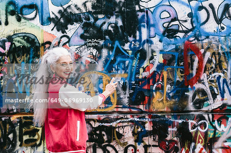 Portrait of mature woman in baseball jacket spray painting graffiti wall