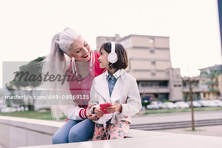 Mature woman and girl sitting on wall listening to headphone music in city