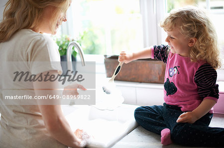 Mother and daughter in kitchen, daughter sitting on work surface helping mother with washing up