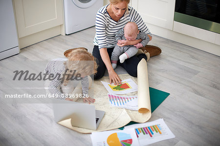 Woman holding baby son in arms, looking through graphs on kitchen floor, young daughter holding paper open