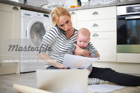 Woman holding baby son in arms, whilst looking through work on kitchen floor, speaking on smartphone and looking at laptop in front of her