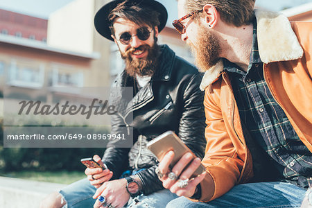 Two young male hipsters sitting on wall with smartphones