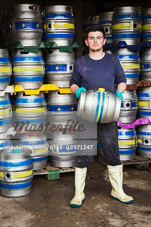 Man working in a brewery, standing next to a stack of metal beer kegs, holding beer keg.