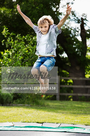 Boy wearing shirt and denim shorts jumping on a trampoline in a garden.