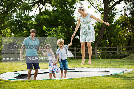 Man, woman, boy and girl holding hands, jumping on a trampoline set in the lawn in a garden.