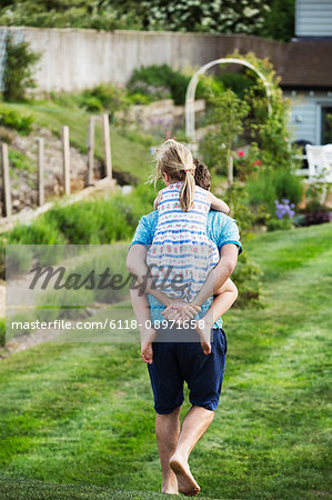 Rear view of a man walking across a lawn, carrying a girl on his back.