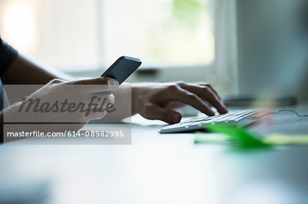 Hand of male designer using smartphone and typing on keyboard at creative studio  desk