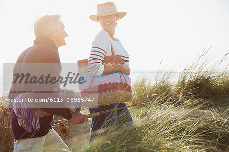 Smiling mature couple with fishing rod walking in sunny beach grass