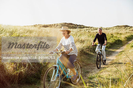 Playful mature couple riding bicycles on sunny beach grass path