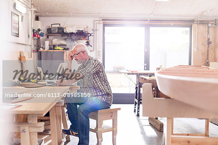 Male carpenter working at laptop on workbench near wood boat in workshop