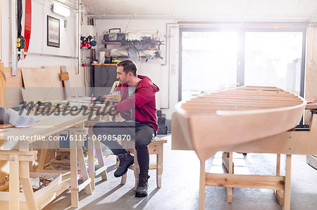 Male carpenter drinking tea and working at laptop on workbench in workshop Male carpenter drinking tea and working at laptop on workbench in workshop