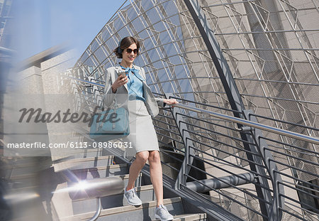 Businesswoman in athletic shoes with cell phone and headphones descending sunny urban stairs