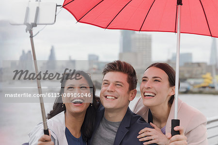 Smiling friend tourists with umbrella taking selfie with selfie stick, London, UK
