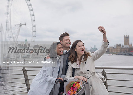 Smiling, happy friends taking selfie with selfie stick on bridge near Millennium Wheel, London, UK