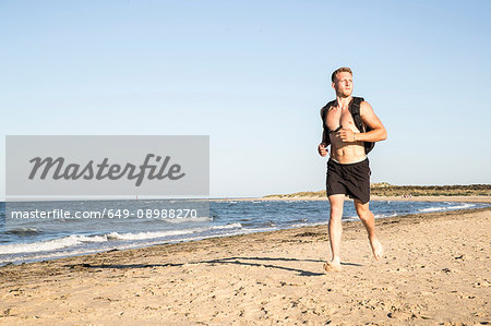 Young male runner wearing shorts and backpack running along beach