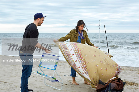 Young sea fishing couple preparing beach chair and picnic blanket on beach