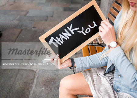 Mid adult sitting outdoors, holding small backboard sign with the words 'Thank you' written in it, mid section