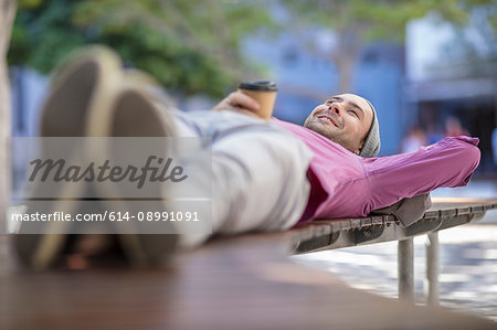 Young man outdoors, lying on bench, holding takeaway coffee cup