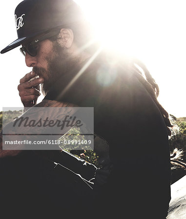 Side view of bearded man wearing baseball cap and sunglasses, smoking cigarette, tattooed arms, sunlight.