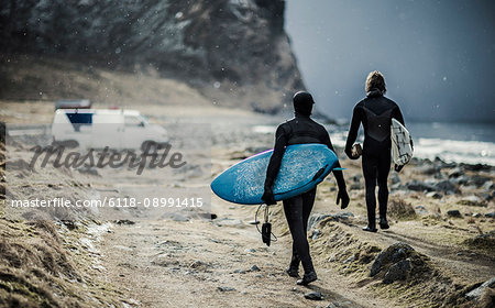 Two surfers wearing wetsuits and carrying surfboards walking towards a van.