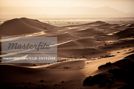 Desert landscape with caravan walking across sand dunes, a plain in the distance.