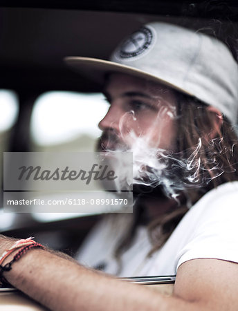 Bearded young man wearing baseball cap sitting in a car, smoking cigarette, cigarette smoke.