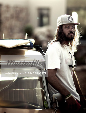 Bearded young man wearing baseball cap leaning against a parked car.