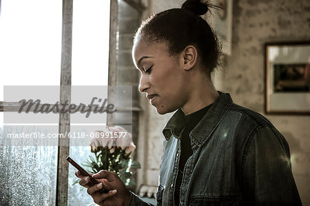 Young woman standing indoors by a window, using cellphone.