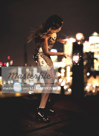 A young woman holding a sparkler and dancing on a building rooftop at night