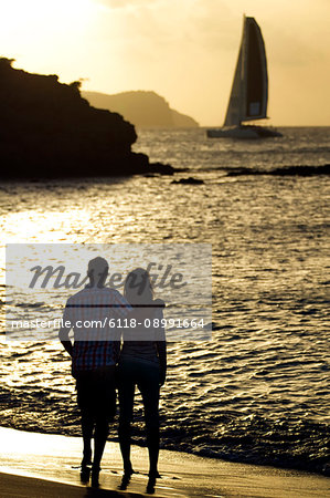 Rear view of couple standing on a sandy beach by the ocean, cliffs and a sailing boat in the distance at sunset.