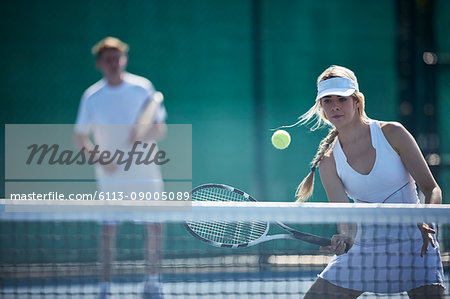 Determined young female tennis player playing tennis, hitting the ball at tennis net on sunny tennis court