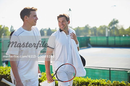 Smiling young male tennis players walking with tennis rackets along sunny tennis court