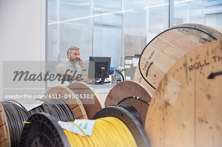 Male supervisor working at computer and talking on ell phone behind spools in fiber optics factory