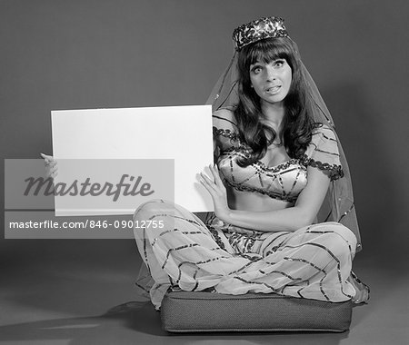 1960s WOMAN IN BELLY DANCE COSTUME SITTING ON CUSHION LOOKING AT CAMERA HOLDING BLANK SIGN LOOKING AT CAMERA