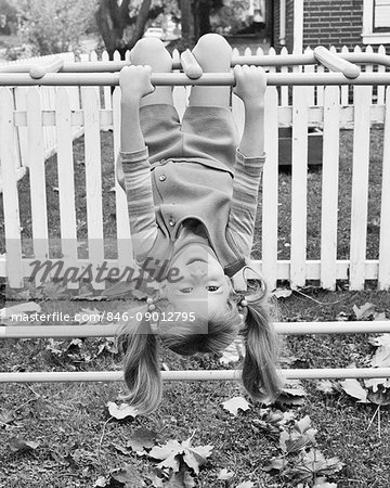 1970s SMILING YOUNG GIRL PIG TAILS HANGING UPSIDE DOWN ON BACKYARD JUNGLE GYM