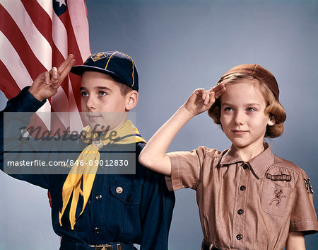 1960s BOY AND GIRL IN CUB SCOUT AND BROWNIE UNIFORMS STANDING BY AMERICAN FLAG SALUTING