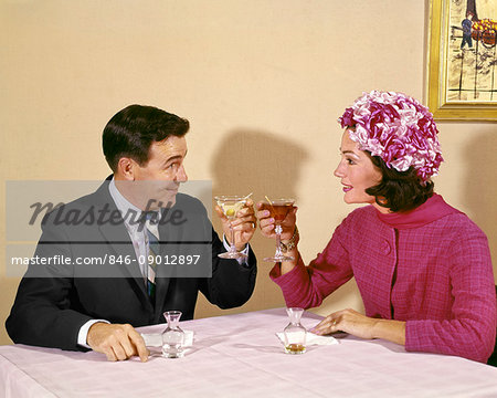 1960s COUPLE TOASTING EACH OTHER WITH WINE IN RESTAURANT WOMAN WEARING FLOWERED HAT